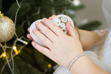 Hands of little girl holding a heart-shaped white Christmas toy near to a decorated Christmas tree.の写真素材