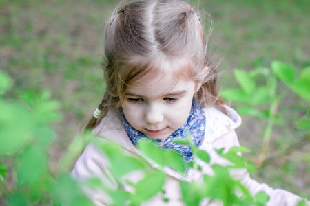 Curious little girl with long blonde hair in pink coat observing something interesting in the grass.