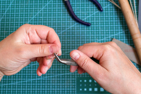 Female hands holding a metal snap hook and tool on a green cutting mat in an jewelry workspace.