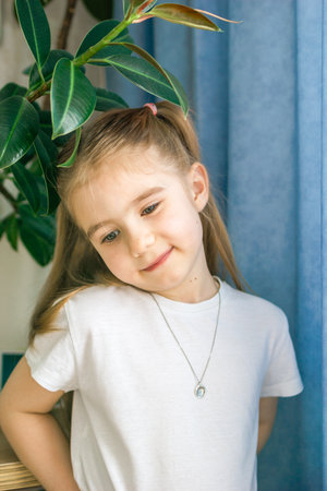 Smiling Little Girl in White Clothes with a Necklace Posing Near Houseplant, Looking at the Camera.の写真素材
