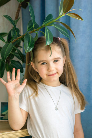 Adorable Little Girl with Brown Hair Waving Hand Surrounded by Green Leaves Indoors.