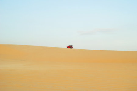 Red Vehicle Driving Across Arid Desert Landscape Under Pale Sky. Mui Ne, Vietnam, White Dunes.の写真素材