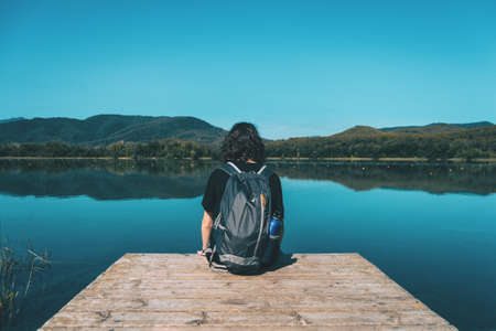 girl sitting on the edge of Banyoles lake on a sunny day with the landscape reflected in the waterの写真素材
