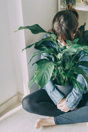 portrait of a woman with brown hair sits on the floor of her house and hugs a plant from a potの写真素材