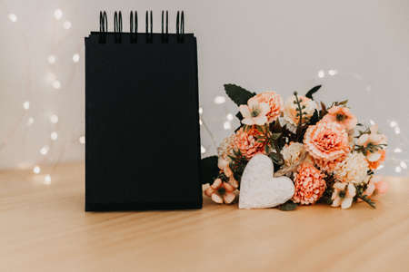 notebook with black sheets with copy space, on a wooden table with a bouquet of flowers and a white heart and bokeh lights around it.の写真素材