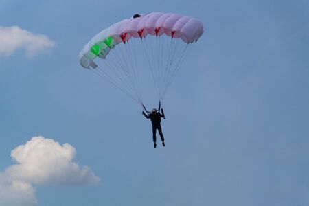 A skydiver in black outfit and black glasses flies across the sky with clouds on a white-red-green...
