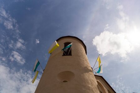 Beige tower of the old catholic church, decorated with white-yellow and white-green flags, against a blue sky with cloudsの写真素材