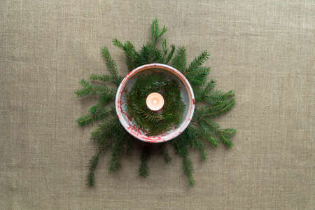 Christmas decor, candlestick-decoration for a New Year's table in the middle of the frame: a burning candle in a plate with water and spruce branches on the background of fir branches and linen canvasの写真素材