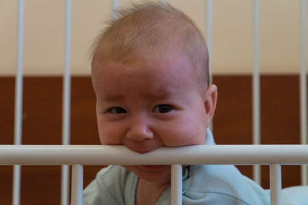 A little child has teeth teething, which is why he gnaws at the edge of a metal bed with his face twisted from pain and discomfortの写真素材