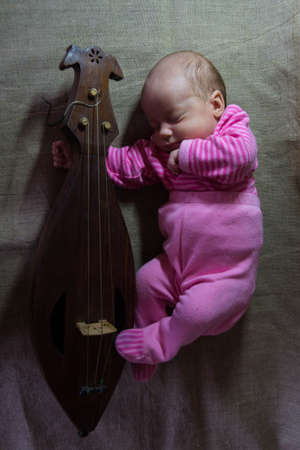 A newborn baby in pink clothes sleeps in an embrace with a wooden old folk stringed musical instrument - a gudok - on a gray linen clothの写真素材