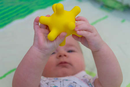 A little child lies on his back and holds a yellow tactile ball with horns in front of him, similar to the coronavirusの写真素材