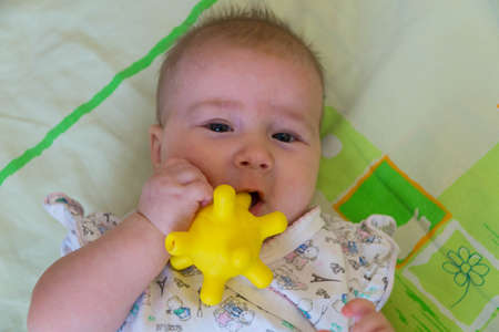 A six-month-old baby lies on his back on a bright blanket and holds in his mouth a yellow toy that looks like a coronavirusの写真素材