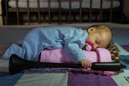 A newborn baby in a blue overalls holds a musical woodwind instrument - a shalmey - in his hand and sleeps on his stomach with a pink pacifier in his mouthの写真素材