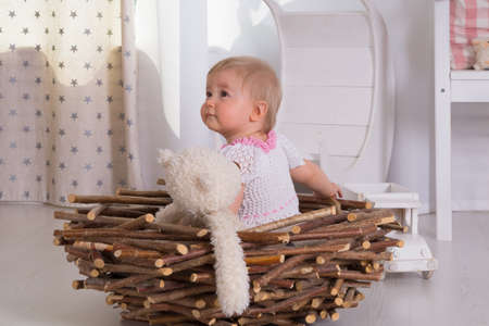 A little one-year-old girl in a white and pink dress sits with a soft toy white bear in a nest of wooden sticks in a room with an interior in light colorsの写真素材