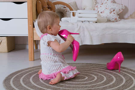 A one-year-old girl in a white and pink openwork dress licks her mother's women's hot pink high-heeled pumps while sitting on the floor in the children's roomの写真素材