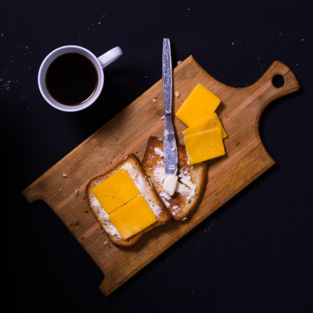 A wooden cutting board holds two slices of toasted bread, spread with butter and topped with cheese, beside it a cup of coffee adds warmth to the kitchen setting.の写真素材