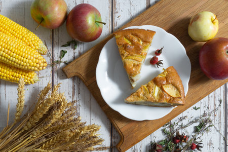 Two slices of apple pie sit on a white plate placed on a wooden board, surrounded by rye ears, corn cobs, fresh apples and sprigs of herbs, all illuminated by soft natural light.の写真素材