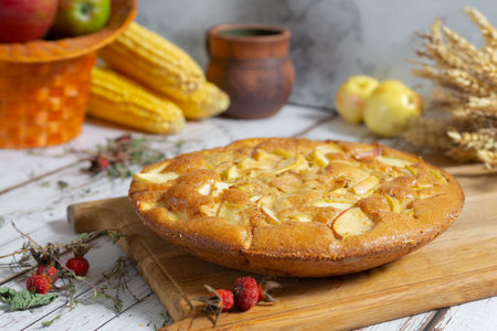 An apple pie sits on a wooden board, surrounded by fresh apples, corn, rose hips and decorative...
