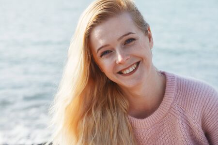 outdoor portrait of pretty blonde young woman in pink knitted sweater looking at camera with...