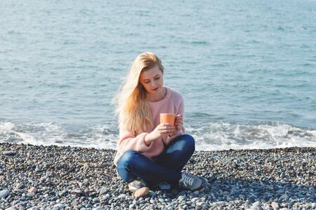 outdoor portrait of pretty blonde young woman in pink knitted sweater sitting at sea shore alone and drinking coffee or teaの写真素材