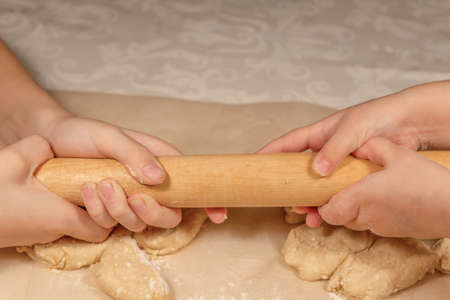 Childrens hands in flour hold a rolling pin against the background of cottage cheese dough on a sheet of baking paperの写真素材