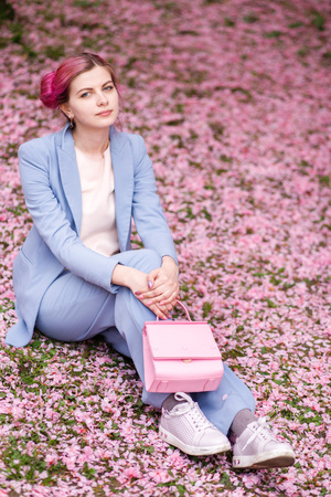 girl with pink hair in a blue stylish suit sitting on a background of pink petals in the gardenの写真素材