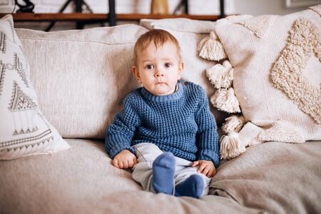 a small boy sits on a sofa in a blue knitted sweater and looks curiously at the camera