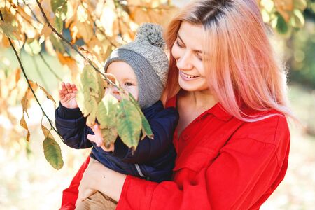 A stylish young mother with a small son in her arms walks in the autumn. Green grass and yellow...