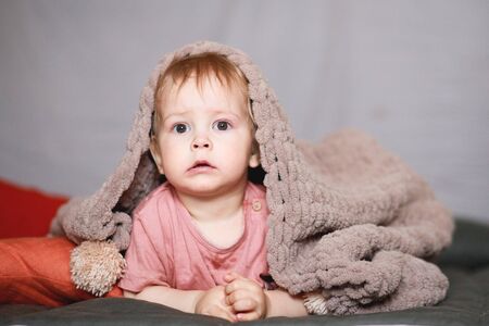a one-year-old boy hides under a beige knitted blanket or bedspread. A boy in bed.の写真素材