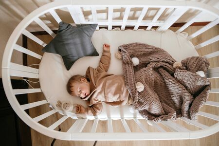 Cute little 1-year-old baby is lying on a cot while sleeping with Windows of light on his face, daytime sleep conceptの写真素材