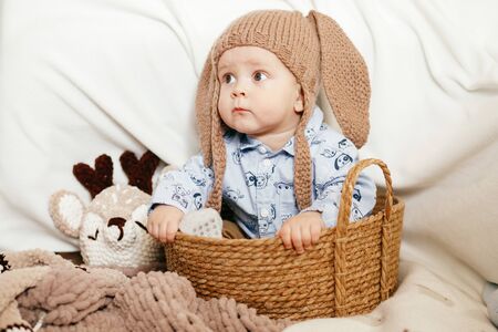 A small boy is sitting in a wicker basket with a hat with rabbit ears on a light background
