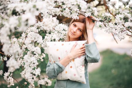 a girl hugs a large pillow with a casual expression on her face on a day off wears sleeping blue pajamas in a blooming Apple orchard in spring.の写真素材