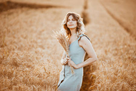 A young girl with long curly hair and a light blue dress, standing in a wheat field, posing for the camera, against the background of the sunsetの写真素材