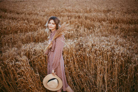 A young woman in a brown dress with white polka dots stands in a wheat field, holding a hatの写真素材