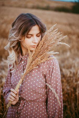 A young woman in a brown dress with white polka dots stands in a wheat field, holding a hatの写真素材