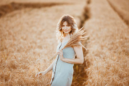 A young girl with long curly hair and a light blue dress, standing in a wheat field, posing for the camera, against the background of the sunsetの写真素材