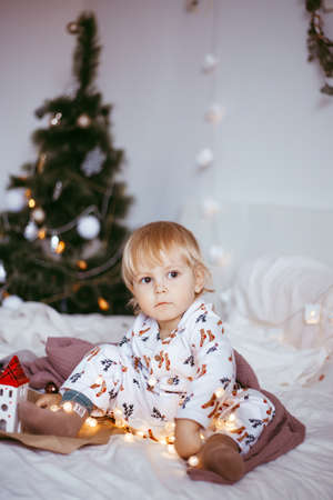 the child's first Christmas. a pensive little boy is lying in his holiday pyjamas on a bed against the background of a brightly decorated fir tree with a Golden light garland. Christmas conceptの写真素材