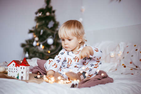 the child's first Christmas. a pensive little boy is lying in his holiday pyjamas on a bed against the background of a brightly decorated fir tree with a Golden light garland. Christmas conceptの写真素材