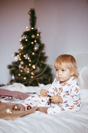 the child's first Christmas. a pensive little boy is lying in his holiday pyjamas on a bed against the background of a brightly decorated fir tree with a Golden light garland. Christmas conceptの写真素材