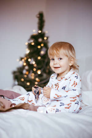 the child's first Christmas. a pensive little boy is lying in his holiday pyjamas on a bed against the background of a brightly decorated fir tree with a Golden light garland. Christmas conceptの写真素材