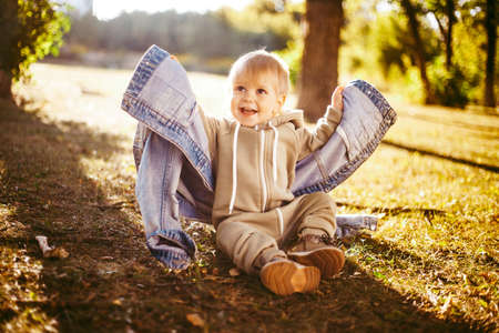 Cute little baby boy in overalls sits and puts on his mother's denim jacket in the autumn Park