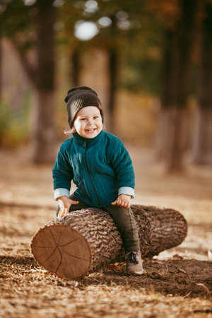 a two-year-old boy in a hat and jacket sits on a log in a Pine forest against the setting sun.の写真素材