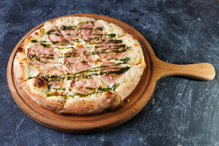 Wooden Board Pizza With Ham, Mozzarella, and Pesto Drizzle on a Dark Surfaceの写真素材