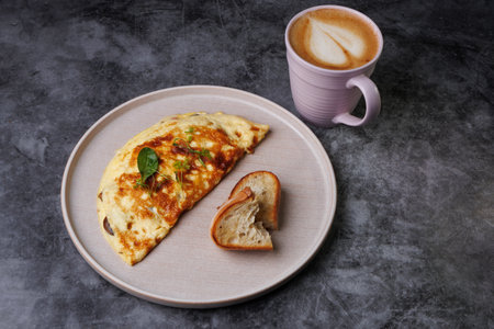 A warm breakfast setting featuring a golden cheese omelette garnished with greens, a slice of crusty bread, served on a pink plate beside a foamy cappuccino. Perfect for food, cafe, morning, and cozy dining themes.の写真素材