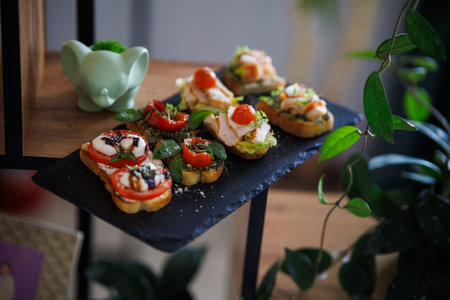An array of bite-sized appetizers arranged on a dark slate platter outdoors. Toasted bread, cherry tomatoes, cheese, radish, greens, and herbs create a vibrant, fresh spread for parties, picnics, or catering.の写真素材