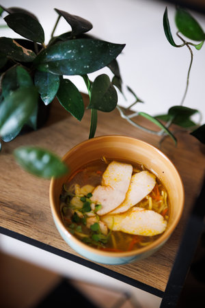 A comforting bowl of herb chicken soup sits on a rustic wooden table, complemented by a white ceramic vase of lavender. Soft focus creates a cozy, homey atmosphere perfect for food and lifestyle.の写真素材