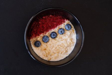 A nourishing bowl of oats topped with blueberries and a splash of red berry powder, presented on a dark ceramic plate to evoke a cozy, healthy breakfast mood.の写真素材