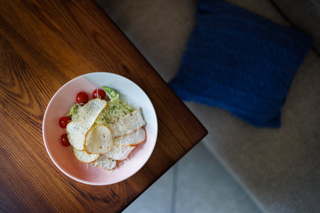A white bowl holds a fresh salad topped with slices of crusty bread, cherry tomatoes, and grated cheese, set on a dark wooden table bathed in warm ambient light for a cozy dining feel.の写真素材