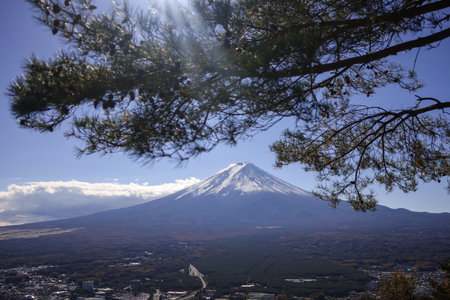 A majestic snow-covered volcano rises above a dark, forested valley. Clear blue sky and scattered clouds frame the peak, evoking rugged nature, travel allure, and alpine scenery suitable for landscape and adventure themes.の写真素材