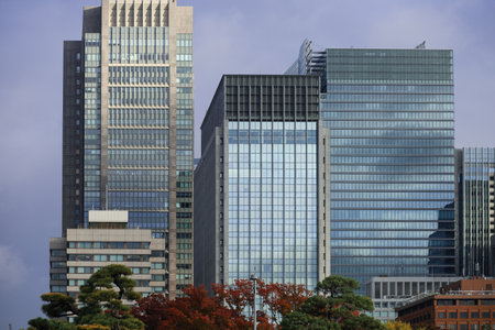 A cluster of modern skyscrapers dominates the skyline, featuring glass facades and steel frames. Foreground trees add color, creating a striking contrast between urban architecture and nature.の写真素材